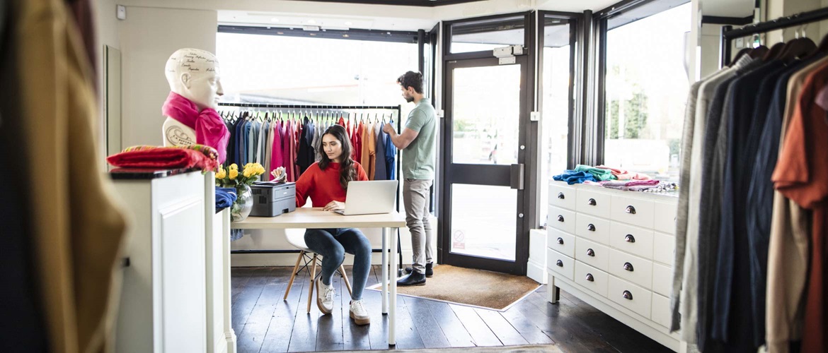 Une femme appartenant au personnel d'un magasin de vêtements est assise sur un bureau pour récupérer un document auprès d'une imprimante Brother, pendant qu'un acheteur de sexe masculin navigue à travers un rail de vêtements derrière elle.